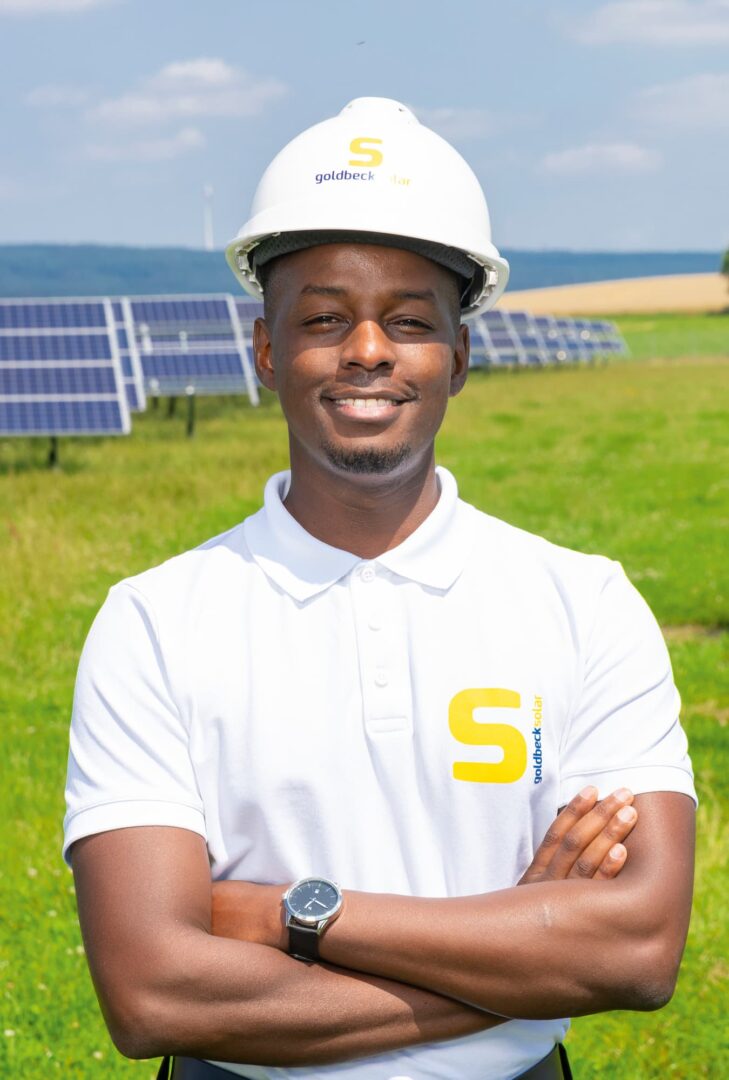 Ein Mitarbeiter von GOLDBECK SOLAR vor einer Reihe Solarpanels einer Photovoltaikanlage auf einer Wiese. Er trägt ein weißes Firmenshirt und einen weißen Helm.